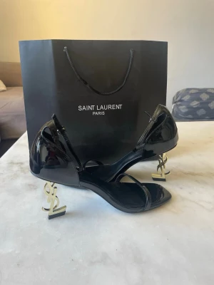 YSL klackar  - Säljer ett par svarta sandaler från Saint Laurent i lackat skinn med öppen tå och smala remmar. Klacken är i metall och formad som YSL-loggan vilket ger en unik och lyxig vibe. Perfekta för dig som vill sticka ut med exklusiv design.