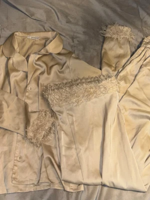 Beige satinpyjamas från Nelly x Glamorous - Snygg beige pyjamas i satin från Nelly x Glamorous. Setet har fluffiga detaljer vid ärmslut och benslut, klassisk krage och knäppning framtill. Materialet är glansigt och mjukt, perfekt för mysiga kvällar hemma.
