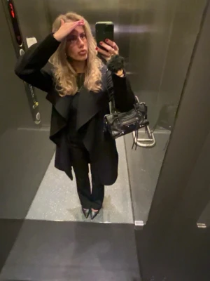 Svart kappa med skärp från Wera - Stilren svart kappa från Wera med bred krage och knytband i midjan. Kappan har en klassisk, rak passform och är tillverkad i ett mjukt material som passar perfekt för kyligare dagar. Enkel att matcha med olika outfits och ger en snygg siluett.