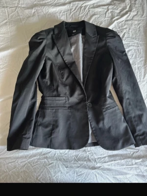 Svart kavaj från H&M - Stilren svart kavaj från H&M med klassisk passform och en knapp framtill. Kavajen har smala slag, två fickor med lock och är helfodrad. Perfekt för en snygg och uppklädd look. Köpt här på Plick men säljer vidare då den var för liten för mig tyvärr..💕