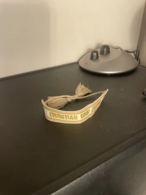 Beige tygarmband Christian Dior - Snyggt beige tygarmband från Christian Dior med texten 'Christian Dior J'Adior' i guld. Armbandet har tofsar i ändarna och är gjort i ett mjukt, vävt material. Perfekt accessoar för dig som gillar exklusiva detaljer och vill sticka ut.