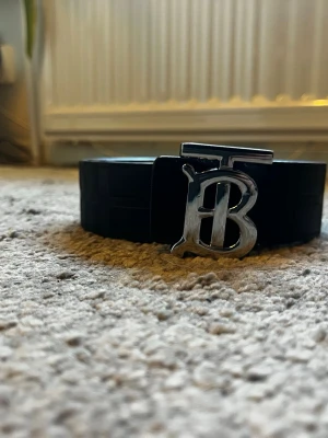 Svart skärp med silverfärgat B-spänne - Snyggt svart skärp från Burberry med ett stort, silverfärgat B-spänne framtill. Skärpet är gjort i skinn och har en stilren, klassisk design som passar till många olika outfits.