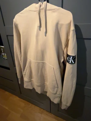 Beige hoodie från Calvin Klein - Snygg beige hoodie från Calvin Klein med klassisk huva och känguruficka. På vänster ärm sitter en svartvit CK-logga som ger en stilren touch. Perfekt för en avslappnad och trendig look.