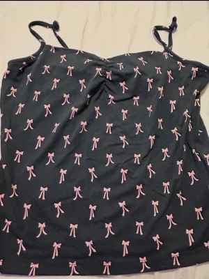 Svart linnetopp med rosa rosetter - Supersöt svart linnetopp med tunna axelband och rosa rosett-mönster över hela. Toppen har en lätt rynkning vid bysten och knyt på axelbanden för extra detalj. Perfekt för dig som gillar gulliga detaljer och en feminin stil.