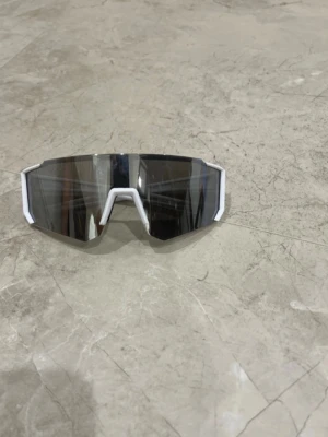 Sportiga vita solglasögon - Säljer ett par sportiga solglasögon med vit båge och stora, spegelblanka svarta linser. De har en futuristisk och kantig design som passar perfekt för en aktiv livsstil. Perfekta för dig som vill sticka ut och skydda ögonen mot solen.