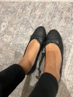 Svarta ballerinaskor  - Stilrena svarta ballerinaskor från C med klassisk rosettdetalj framtill och präglad logotyp på tån. Skorna är tillverkade i skinn och har en elegant, tidlös design som passar perfekt till många olika outfits.