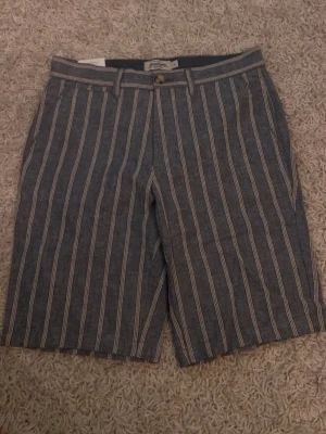 Randiga linneshorts från Dressmann - Säljer dessa helt nya och oanvända shorts från Dressmann i skönt linnematerial. Shortsen har normal passform, bälteshällor och knappar bak. Perfekta för varma dagar och ger en avslappnad men stilren look.