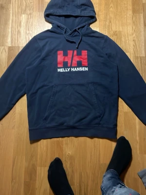 Mörkblå Helly Hansen hoodie S - Säljer en mörkblå hoodie från Helly Hansen med stor röd HH-logga och vit text på bröstet. Klassisk känguruficka framtill och justerbar huva med dragsko. Perfekt för dig som gillar streetwear och vill ha något bekvämt och stilrent.