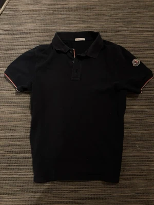 Svart pikétröja från Moncler - Säljer en snygg Piké från Moncler! Piken är i mycket bra skick och har inga defekter! Knappt använd. Storlek M. Qr-koden funkar som den ska. Om ni har några frågor så är de bara att höra av er!😊