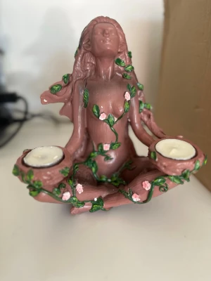 Gudinneliknande ljushållare staty - Unik staty i form av en sittande gudinna med blommor och blad slingrande runt kroppen. Figuren är brun med gröna och rosa detaljer och håller två ljushållare i händerna. Perfekt som dekoration eller för att skapa mysig stämning med värmeljus.