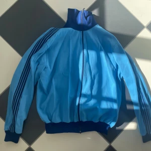 Blå Vintage tracksuit jacka - Snygg retro träningsjacka i ljusblått med mörkblå detaljer och dragkedja framtill. Jackan har hög krage, muddar vid ärmslut och midja samt tre mörkblå ränder längs ärmarna. Trendig och supersnygg! Passar perfekt nu till vår och sommar 