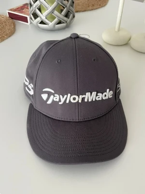 Taylormade Stealth golfkeps grå - Snygg mörkblå keps från TaylorMade med böjd skärm och vit broderad logga framtill och STEALTH texten på sidan. Klassisk sexpanelsdesign med ventilationshål och justerbar baksida. Perfekt för golfbanan!