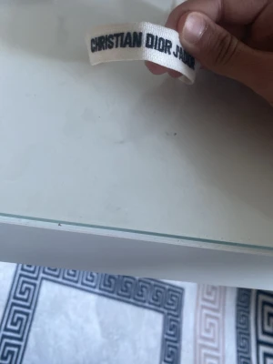 Vitt Christian Dior tygarmband - Snyggt vitt armband från Christian Dior i tyg med svart broderad text. Armbandet har justerbar knytning och dekorativa tofsar i ändarna. Perfekt accessoar för att lyfta din outfit med en exklusiv touch.