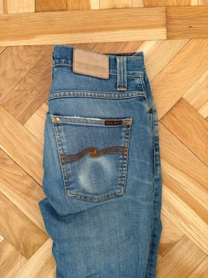 Blå jeans från Nudie Jeans - Säljer ett par klassiska blå jeans från Nudie Jeans med snygg slitning och kontrastsömmar. Modellen har fem fickor och en rak passform. Materialet är mjuk denim i bomull och bakfickan har Nudie Jeans ikoniska brodyr.