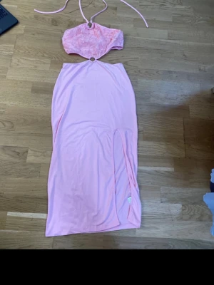 Ljusrosa långklänning med slits - Trendig ljusrosa långklänning med halterneck och öppen mage. Överdelen har 3D-blommor och ringdetalj framtill. Kjolen har hög slits och mjukt fall. Perfekt för dig som vill sticka ut med en unik och feminin look.