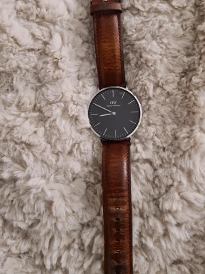 Daniel Wellington klocka med brunt läderband - Stilren klocka från Daniel Wellington med svart urtavla och silverfärgad boett. Klockan har ett klassiskt brunt läderband och minimalistisk design. Perfekt för dig som gillar enkel och tidlös stil.