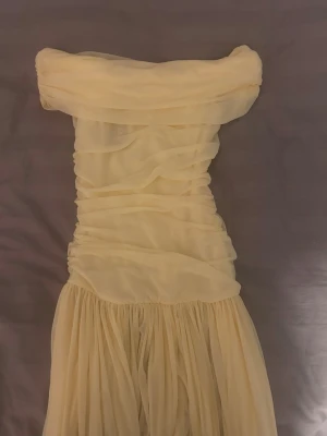 Ljusgul axelbandslös långklänning - Helt ny aldrig använd (prislappen kvar). Superfin ljusgul långklänning med axelbandslös design och draperad överdel. Klänningen har ett flowy, plisserat kjolparti och är tillverkad i ett tunt, skirt material som ger en romantisk känsla.