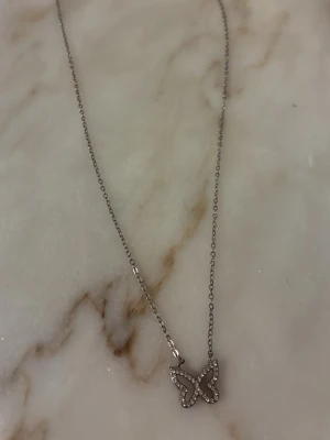 Silverfärgat halsband med fjärilshänge - Snyggt halsband i silverfärg med ett fjärilsformat hänge dekorerat med små pärlor. Kedjan är tunn och elegant, perfekt för att ge en söt touch till din outfit. Fjärilshänget har en modern design och pärldetaljerna ger extra glans.