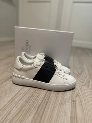 Valentino Garavani vita sneakers - Snygga vita sneakers från Valentino Garavani med svart band över sidan och coola nitar på hälen. Skorna har en klassisk låg siluett och är tillverkade i slätt läder med vita snören och diskret logga. Perfekta för dig som gillar stilrena och exklusiva sneakers. Dem ser lite svarta ut men dem är marinblåa. Allt i bilden får man med.