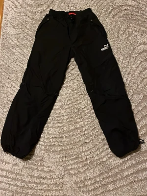 Svarta mjukisbyxor från Puma - Svarta mjukisbyxor från Puma med logga på låret. Byxorna har elastisk midja, snörning vid benslut och praktiska fickor. Perfekta för chill eller träning, tillverkade i ett lätt och bekvämt material.