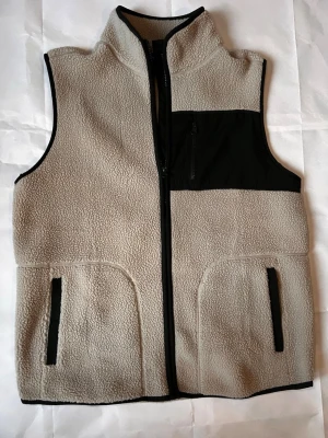 Beige fleeceväst från Newbody - Beige fleeceväst med svarta detaljer från Newook. Västen har hög krage, hel dragkedja framtill, två fickor med svarta kanter och en svart bröstficka med dragkedja. Perfekt för lager-på-lager och chill stil.