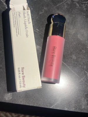 Rare Beauty Soft Pinch Dewy Liquid Blush - Flytande rouge från Rare Beauty i nyansen 'Hope', en mjuk rosa färg. Kommer i en stilren frostad glasflaska med guldig och silverfärgad kork. Perfekt för att ge kinderna en fräsch och naturlig lyster.