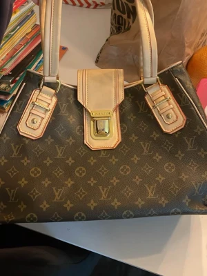  handväska - Säljer en klassisk handväska  monogrammönster med beigea detaljer och guldfärgade metalldelar. Väskan har två handtag och ett lock med spänne framtill. Väskan är rymlig, lite sprucken i tyget se bild 2 