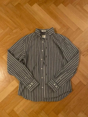 Randig blå skjorta med knappar Gant - Snygg blå skjorta från Oscar Jacobson med vita vertikala ränder och klassisk krage. Skjortan har långa ärmar, knappar framtill och vid manschetterna. Tillverkad i mjuk bomull som känns skön mot huden. Perfekt för en stilren och avslappnad look. Man