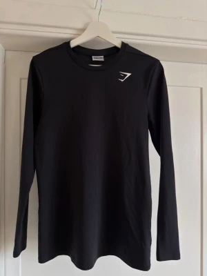 Svart långärmad träningströja Gymshark - Snygg svart långärmad träningströja från Gymshark med rund hals och diskret logga på bröstet. Tillverkad i ett mjukt material som passar perfekt för gymmet eller löprundan.