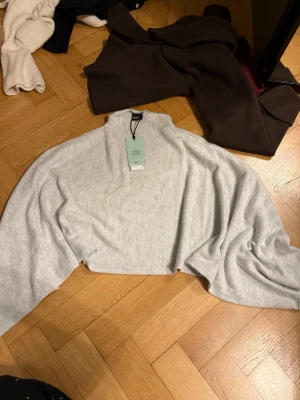 Ljusgrå cape från Gina Tricot - Mjuk och stilren ljusgrå cape från Gina Tricot i färgen grey melange. Capen har hög krage och vida ärmar, perfekt för lager på lager och en chill look. Materialet känns skönt mot huden och ger en avslappnad vibe.