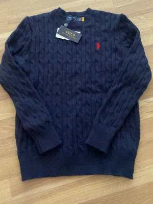 Mörkblå kabelstickad tröja Polo Ralph Lauren - Snygg mörkblå kabelstickad tröja från Polo Ralph Lauren med klassisk röd logga broderad på bröstet. Tröjan har rund halsringning, långa ärmar och ribbade muddar. Perfekt för dig som gillar tidlös och stilren design.