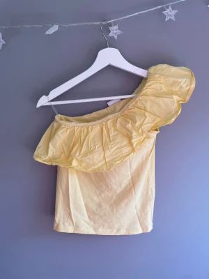Gul oneshoulder topp med volang - Snygg ljusgul oneshoulder topp med stor volang längs halsen. Perfekt för dig som vill sticka ut med en unik och trendig stil. Toppens material är mjukt och lätt, vilket gör den bekväm att bära under varma dagar. passar som en XS