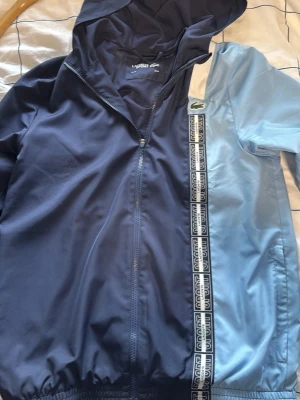 Tvåfärgad vindjacka från Lacoste - Snygg vindjacka från Lacoste i två nyanser av blått, med huva och dragkedja. Ena sidan är mörkblå och den andra ljusblå, med vita detaljer och Lacoste-logga på bröstet. Jackan har sportiga band med texten 'Lacoste Sport' längs framsidan.