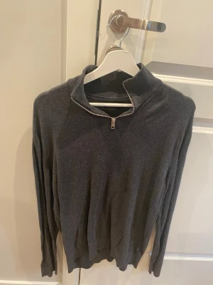 Mörkgrå halvzip tröja - Mörkgrå långärmad tröja med halv dragkedja i halsen. Tröjan har en enkel och stilren design med ribbade muddar vid ärmslut och nederkant. Perfekt för lager på lager och passar till det mesta.