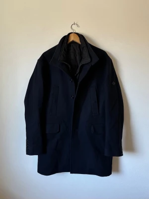 Mörkblå klassisk rock från J.Lindeberg - Stilren mörkblå rock med avtagbar väst från J.Lindeberg med rak passform och knäppning framtill. Rocken har två sidofickor med lock och en hög krage som kan stängas med knappar. Materialet är kraftigt och passar perfekt för kyliga dagar. 6R17