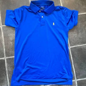 Blå pikéskjorta från Polo Ralph Lauren - Säljer en klassisk blå pikéskjorta från Polo Ralph Lauren med gul broderad logga på bröstet. Skjortan har korta ärmar, krage och knappar framtill. Perfekt för dig som vill ha en stilren och tidlös look. Pris ej hugget i sten, postar inom 1-2 dagar.