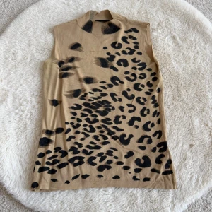 Leopardmönstrad polotopp från Gianini (S) - Cool beige polotopp från Gianini med svart leopardmönster. Toppen är ärmlös och har en ribbad polokrage som ger en snygg och trendig look. Perfekt för dig som gillar att sticka ut och vill ha något unikt i garderoben.