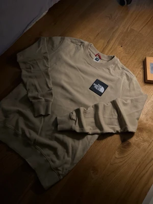 Beige sweatshirt från The North Face - Snygg beige sweatshirt från The North Face med rund halsringning och ribbade muddar. Stor logga i svart och vitt på bröstet ger en clean och modern look. Perfekt till jeans eller joggers för en avslappnad stil.