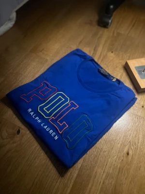 Blå POLO Ralph Lauren t-shirt - Snygg blå t-shirt från POLO Ralph Lauren med stort broderat POLO-tryck i flera färger på bröstet. Klassisk rund hals och korta ärmar. Tillverkad i mjuk bomull som känns skön mot huden. Perfekt för en avslappnad och trendig look.