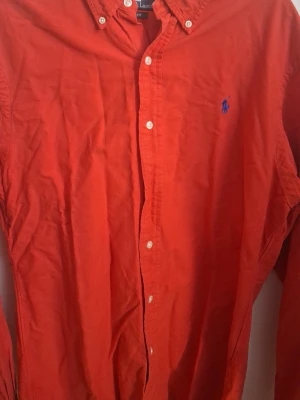 Röd skjorta från Polo Ralph Lauren - Säljer en röde långärmad skjorta från Polo Ralph Lauren med slim fit-passning. Skjortan har klassisk krage, knappar framtill och blå broderad logga på bröstet. Perfekt för dig som vill ha en färgstark och stilren look.