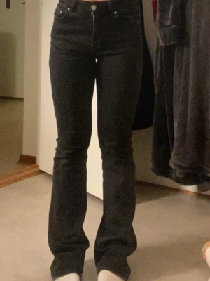 Svarta bootcut jeans med hög midja - Säljer ett par svarta bootcut jeans med hög midja och klassisk femficksdesign. Jeansen har en snygg passform som sitter tight upptill och släpper ut vid benen. Materialet är denim och de har knapp- och dragkedjeförslutning.