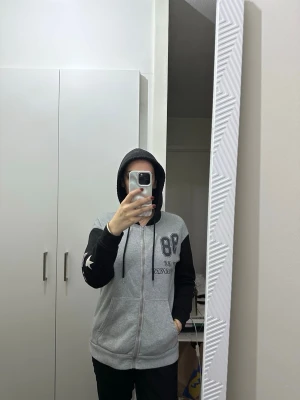 Grå och svart hoodie med tryck - Säljer en grå hoodie med svarta ärmar och huva. Dragkedja framtill, fickor på sidorna och tryck med '98' och 'Colorado' på ryggen samt stjärna på ena ärmen. Skön och avslappnad passform, perfekt till vardags. Materialet känns mjukt och bekvämt.