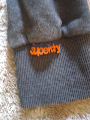 Mörkgrå hoodie från Superdry - Snygg mörkgrå hoodie från Superdry med stora tryck i rött och blått framtill samt orange broderad logga vid ärmslutet. Hoodien har ribbade muddar och en logga på axeln den har en snygg relaxed slim fit look.