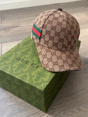 Gucci GG Supreme keps brun - Exklusiv keps från Gucci i GG Supreme-mönster, brun med klassiska gröna och röda ränder på sidan. Justerbar rem baktill och detaljerad logotyp på insidan. Materialet är canvas med läderdetaljer. Perfekt för dig som vill ha en lyxig och trendig accessoar.