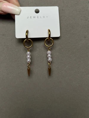 Guldörhängen  - Snygga örhängen i guldfärgad metall med tre vita pärlor på varje hängande del och en spetsig detalj längst ner. Perfekt för dig som gillar trendiga accessoarer med en cool touch. Örhängena har rund ring och stilren design.