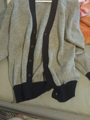 Grå kofta från Comme des Garçons - Snygg grå kofta från Comme des Garçons Homme med mörkblå knappar och kantdetaljer. Koftan har ribbstickade muddar och klassisk v-ringning. Perfekt för lager-på-lager och stilrena outfits.