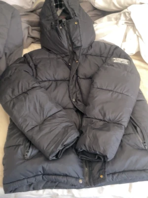 Burberry England svart pufferjacka - Svart pufferjacka från Burberry England med avtagbara ärmar och huva. Jackan har ett rutigt innerfoder och logotyp på ena ärmen. Quiltad nylon och praktiska fickor framtill. Kan användas både som jacka och väst för en flexibel look.