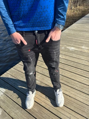 Svarta distressed jeans med färgstänk - Säljer ett par svarta jeans med slim fit och slitningar på knäna. Jeansen har vita och röda färgstänk för en unik streetwear-look. Klassisk femficksmodell och knappgylf. Perfekta för dig som gillar en edgy stil.