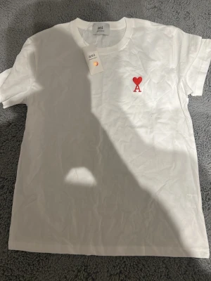 Vit t-shirt från Ami  - Vit t-shirt från Ami med klassisk passform och korta ärmar. På bröstet finns en broderad röd hjärta och bokstaven A. Materialet är mjuk bomull och t-shirten har en rund halsringning. Perfekt för en clean och stilren look.