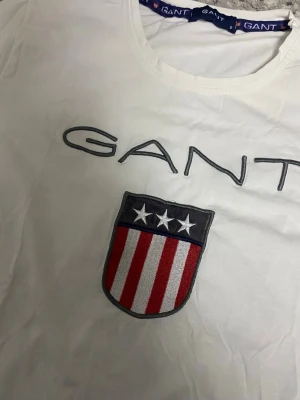 Vit GANT t-shirt med emblem - Vit t-shirt från GANT med stort broderat emblem på bröstet i rött, vitt och blått samt GANT-logga ovanför. Klassisk rund hals och korta ärmar. Tillverkad i mjuk bomull för en skön känsla. Perfekt för en clean och sportig look.
